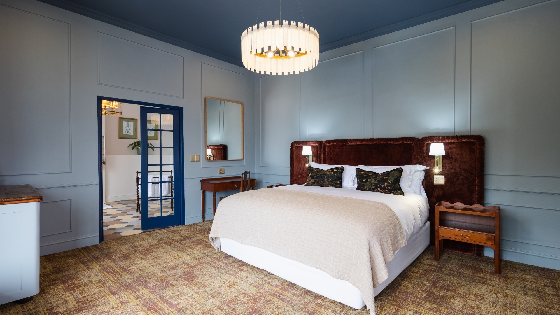 Zomerlust Boutique Hotel Paarl. Elegant bedroom featuring slate blue wainscoted walls, a deep blue ceiling, a plush brown velvet headboard on a king-size bed, and a modern tiered chandelier.