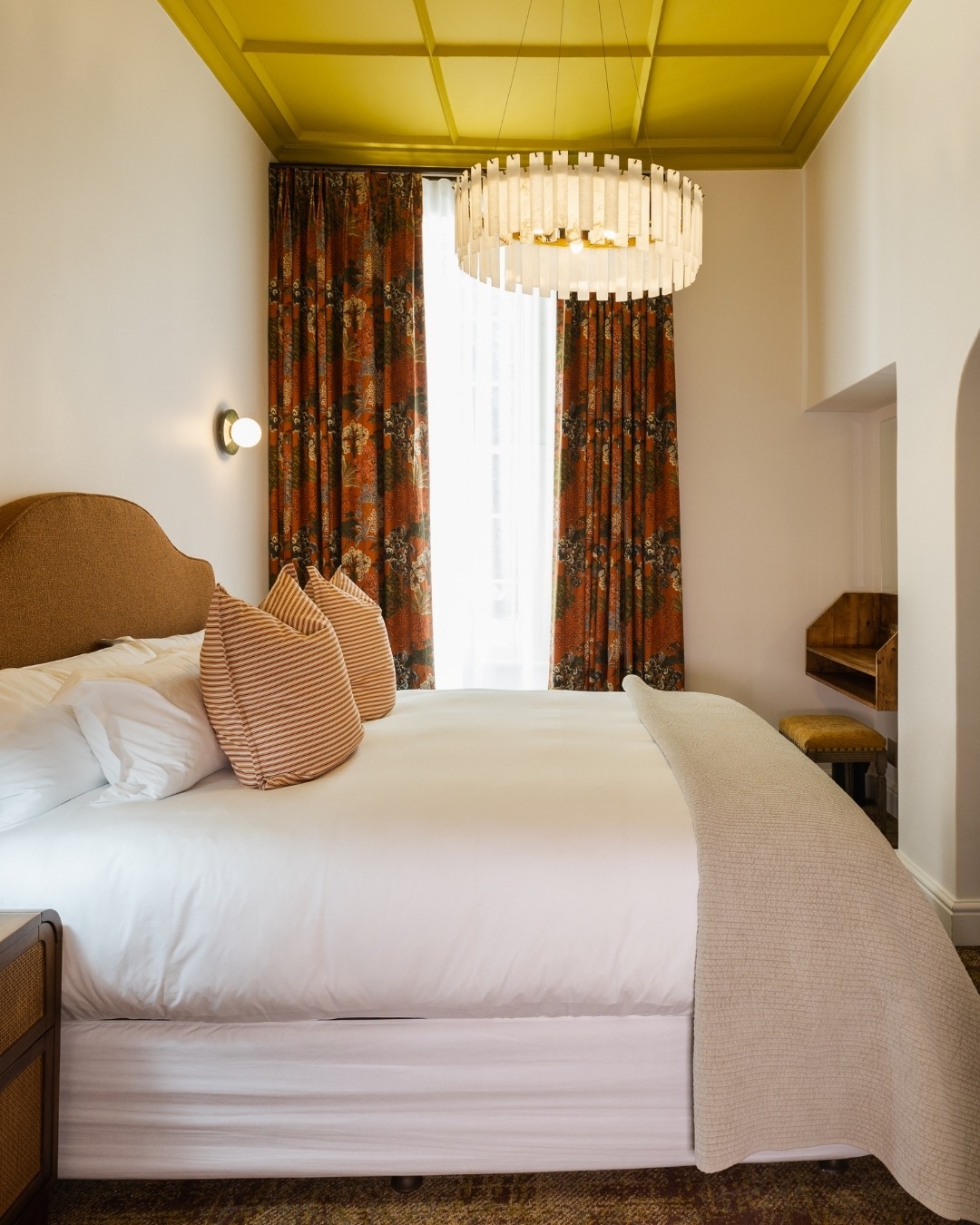 Zomerlust Boutique Hotel Paarl. Bright bedroom with a bold mustard-yellow coffered ceiling, a circular frosted-glass chandelier, patterned drapes framing a window, and a bed with striped throw pillows.